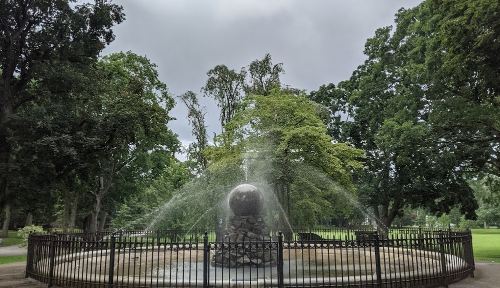 Stone fountain at Capron Park in Attleboro, MA, with water jets arching upward, surrounded by lush greenery, enhancing the park's tranquil ambiance.