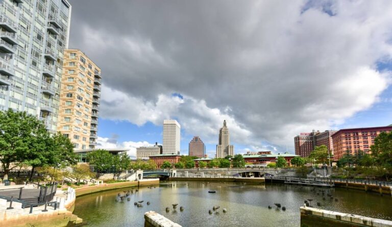 Scenic view of Waterplace Park in Providence, Rhode Island, featuring the river, arched pedestrian bridges, and a waterfront walkway lined with trees and historic buildings.