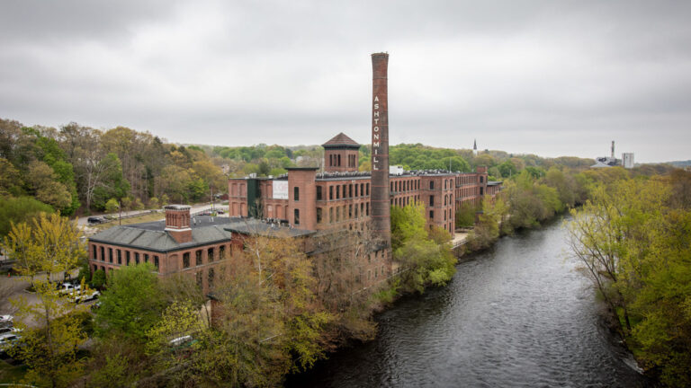 Aerial view of Ashton Mill alongside the Blackstone River in Cumberland, RI, with iconic brick architecture and green surroundings.